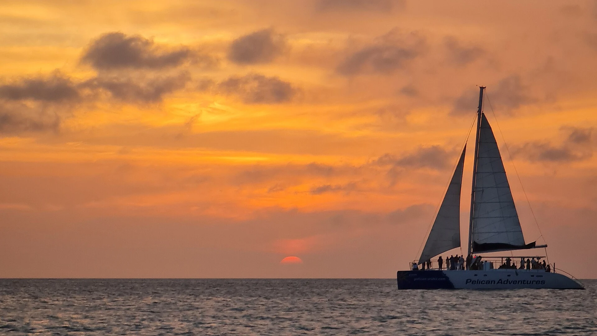 A sailing catamaran of passengers silhouetted against an orange sunset over the sea