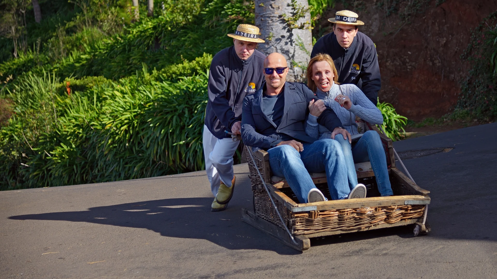 Two straw-hatted carreiros steering a wicker toboggan with passengers down a street in Monte, Funchal